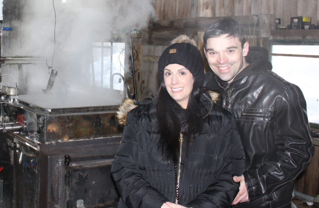 Young couple standing in a maple sugar house with steam from the syrup boiler billowing behind them.