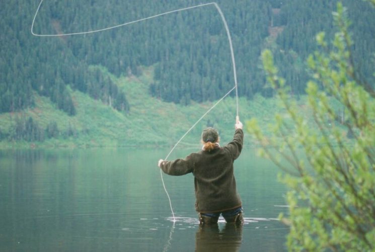 Woman in brown coat standing in river fly fishing with tree-covered mountain in the background