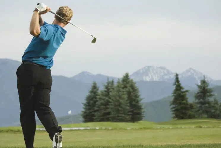 Man in blue shirt and black pants swinging a golf club on a golf course with mountains in the background