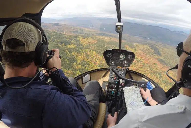 View from inside of a helicopter flying over mountains with green, yellow, and orange trees