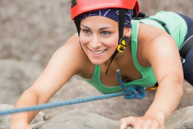 Close up view of a woman rock climbing dressed in a green tank top, navy pants, and red helmet