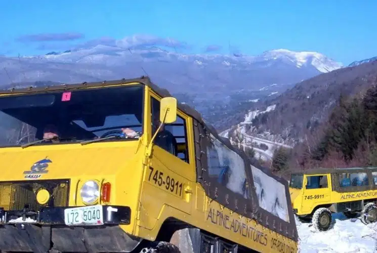 Two yellow off-road tour trucks driving up a snow covered trail with mountains in the background