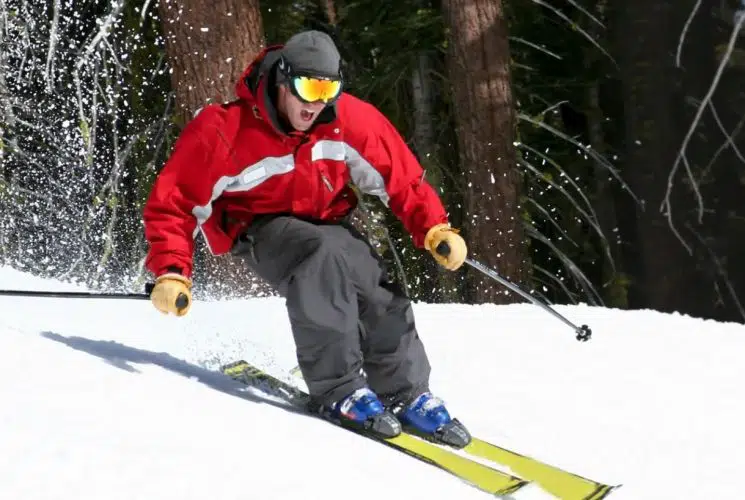 Man in a red coat and gray pants with yellow skis snow skiing down a mountain