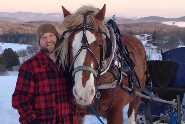 Large brown and white horse pulling a blue sled in the snow with a man wearing a red and black coat and brown hat standing next to the horse