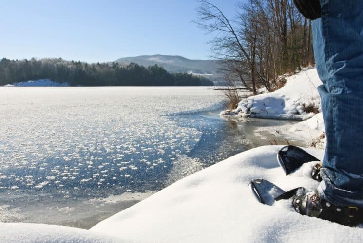 Close up view of a person's legs wearing jeans and snowshoes in the snow standing near a frozen body of water