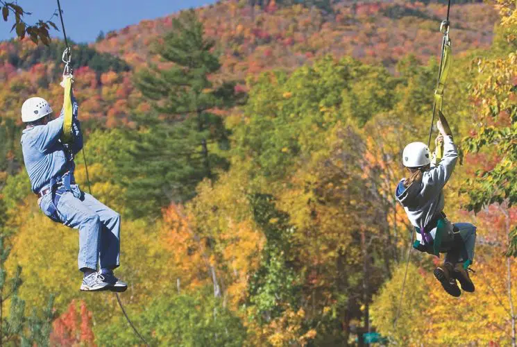 Two people ziplining over many trees with green, yellow, and orange leaves