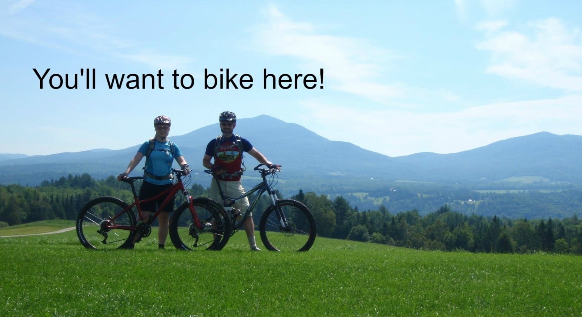 two people standing next to their mountain bikes on a grassy hill.