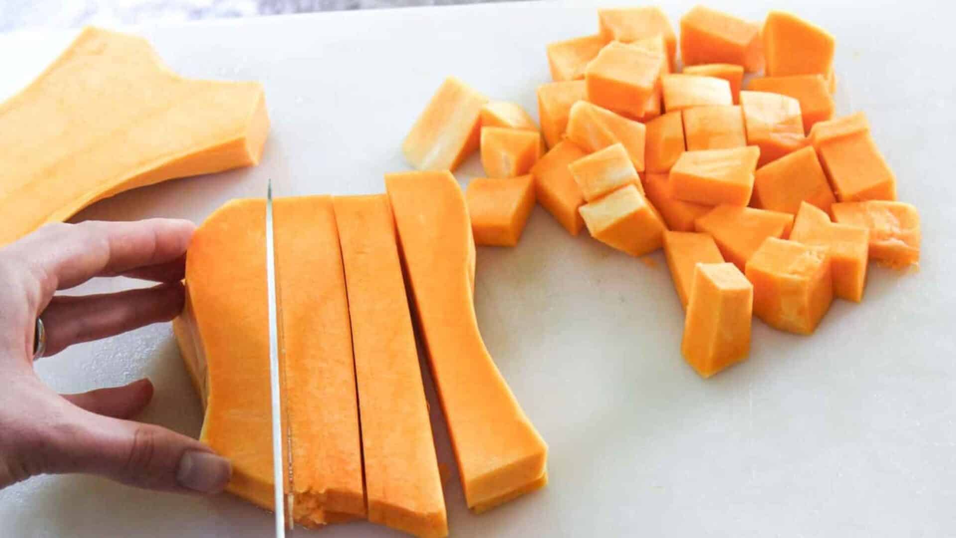 hand holding knife cutting butternut squash into cubes on a cutting board