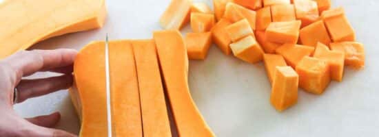 hand holding knife cutting butternut squash into cubes on a cutting board