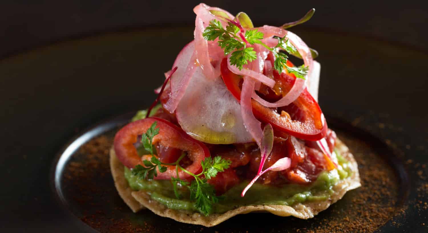 Close up view of a beef tartar tostada topped with red peppers and red onions on a black porcelain plate
