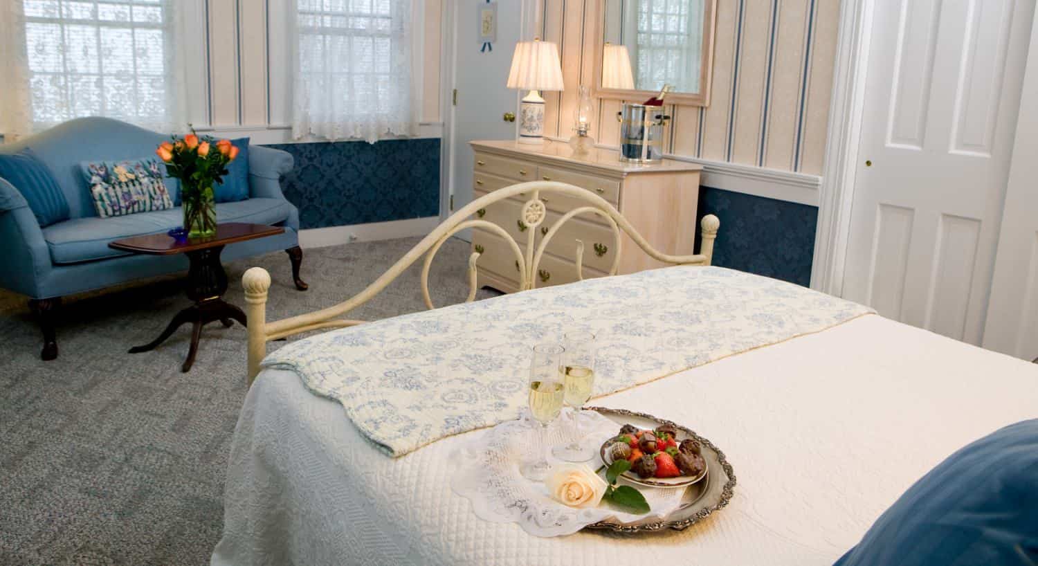 Bedroom suite with gray carpeting, cream and blue striped wallpaper, blue brocade wallpaper below chair rail, cream wrought iron bed, white bedding, and sitting area