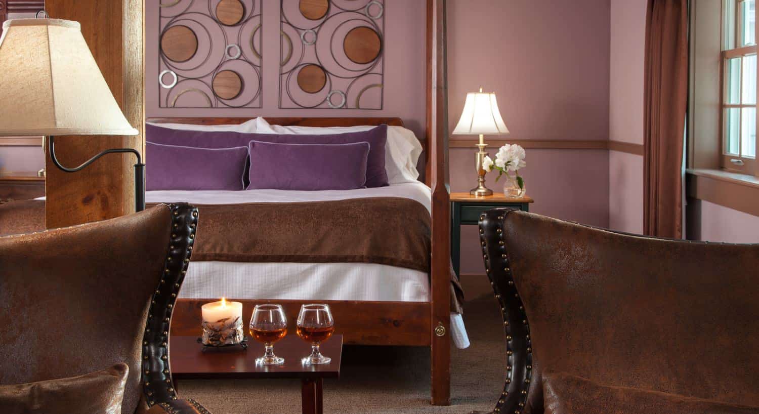 Large bedroom suite with dark brown carpet, light lavendar walls, dark wooden four-poster bed, white bedding, purple pillows, and sitting area