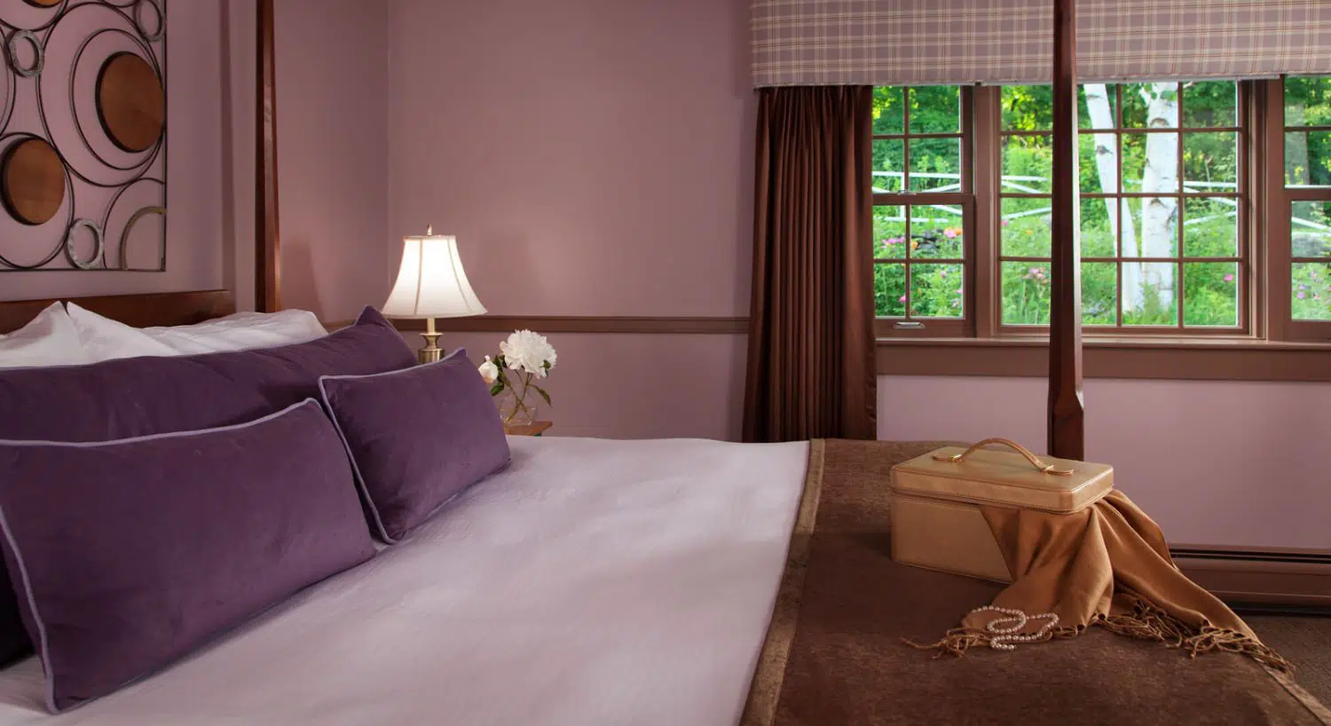 Bedroom with dark wooden four-poster bed, white bedding, purple pillows, brown blanket, and lavendar walls