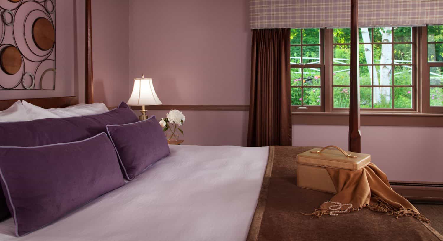 Bedroom with dark wooden four-poster bed, white bedding, purple pillows, brown blanket, and lavendar walls