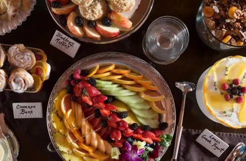 Close up view of a dark wooden table with multiple dishes filled with pastries, muffins, yogurt, and sliced fruit
