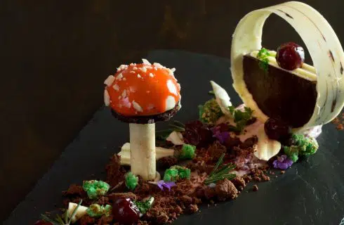 Close up view of a fancy dessert dish on black slate tile which looks like a orange mushroom surrounded by crumbles next to a ring of white and dark chocolate