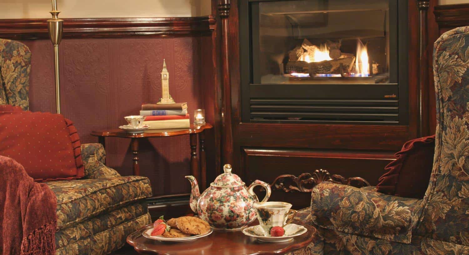 Close up view of sitting area with dark floral upholstered arm chairs, dark wooden coffee table, antique tea pot, tea cup and saucer, plate of cookies, and a fireplace