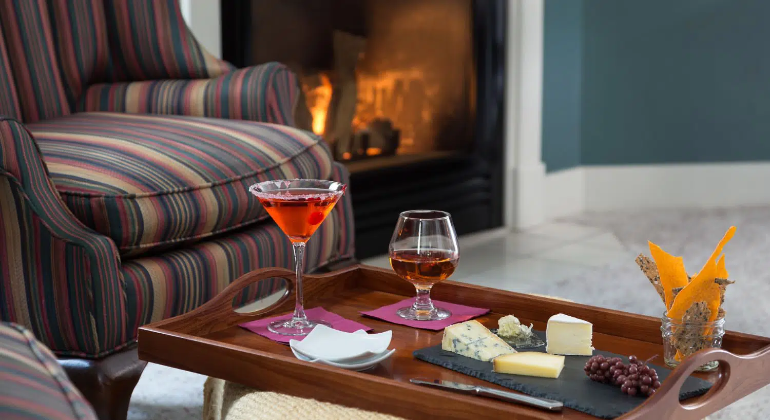 Close up view of wooden tray with cocktails, cheeses, crackers, and fruit next to a striped upholstered arm chair and fireplace