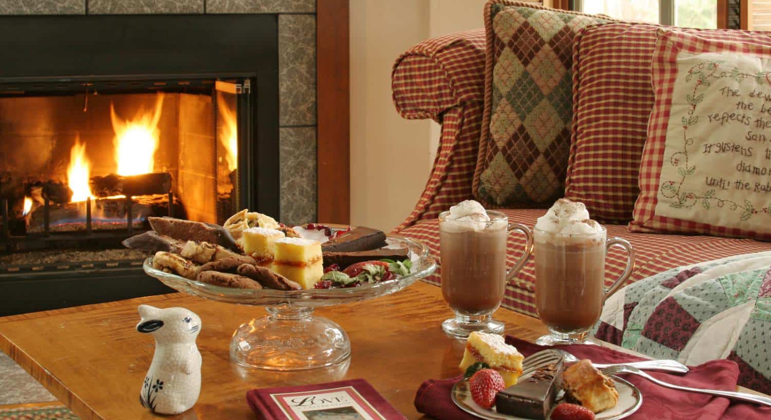 Close up view of glasses full of hot chocolate and serving dish full of desserts on wooden coffee table next to red-checked upholstered couch and fireplace