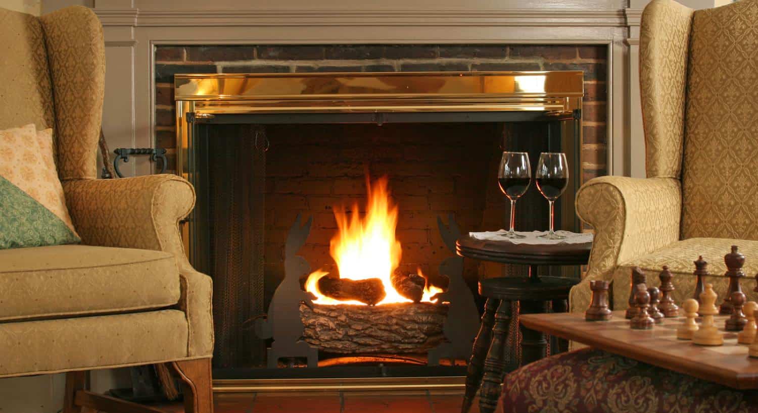 Close up view of sitting area with light-tan upholstered arm chairs, dark wooden stool, glasses full of wine, and a fireplace