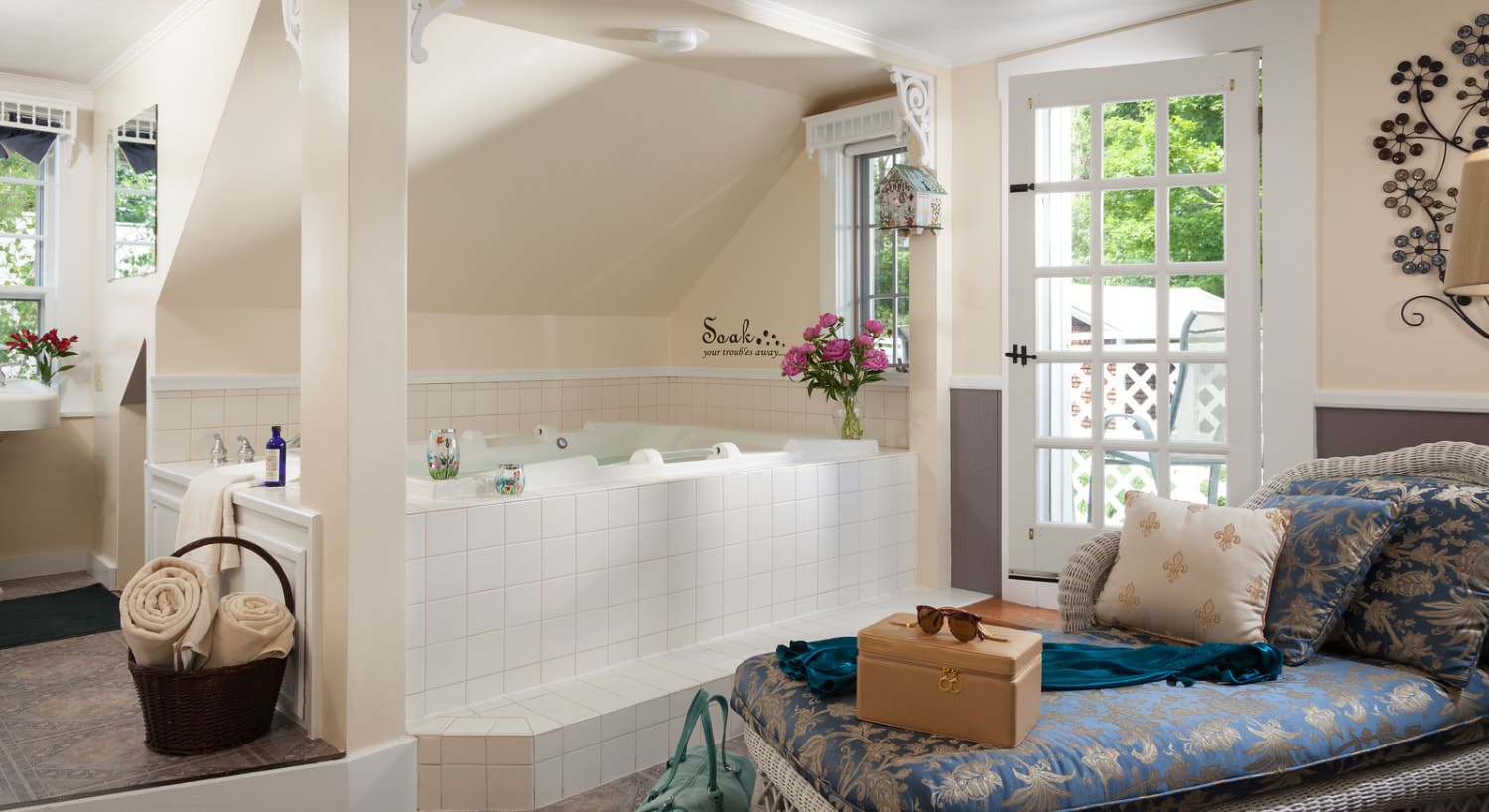 Large whirlpool tub surrounded by white tiles and light cream walls, next to a door to the balcony, and white wicker chaise with blue and gray floral cushions