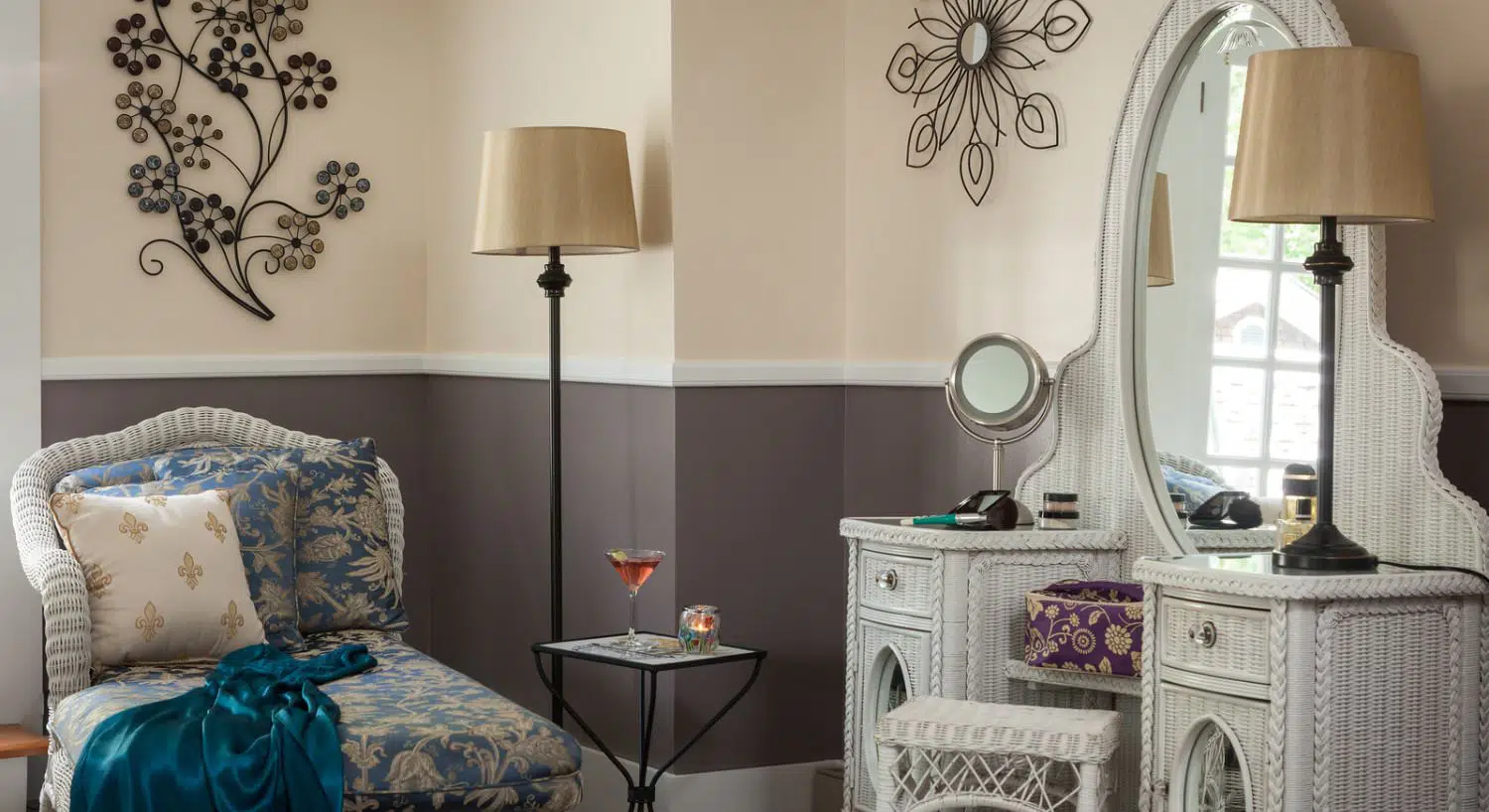 Room with cream walls on top and dark gray walls below chair rail, white wicker chaise and make-up dresser, and wrought iron lamps