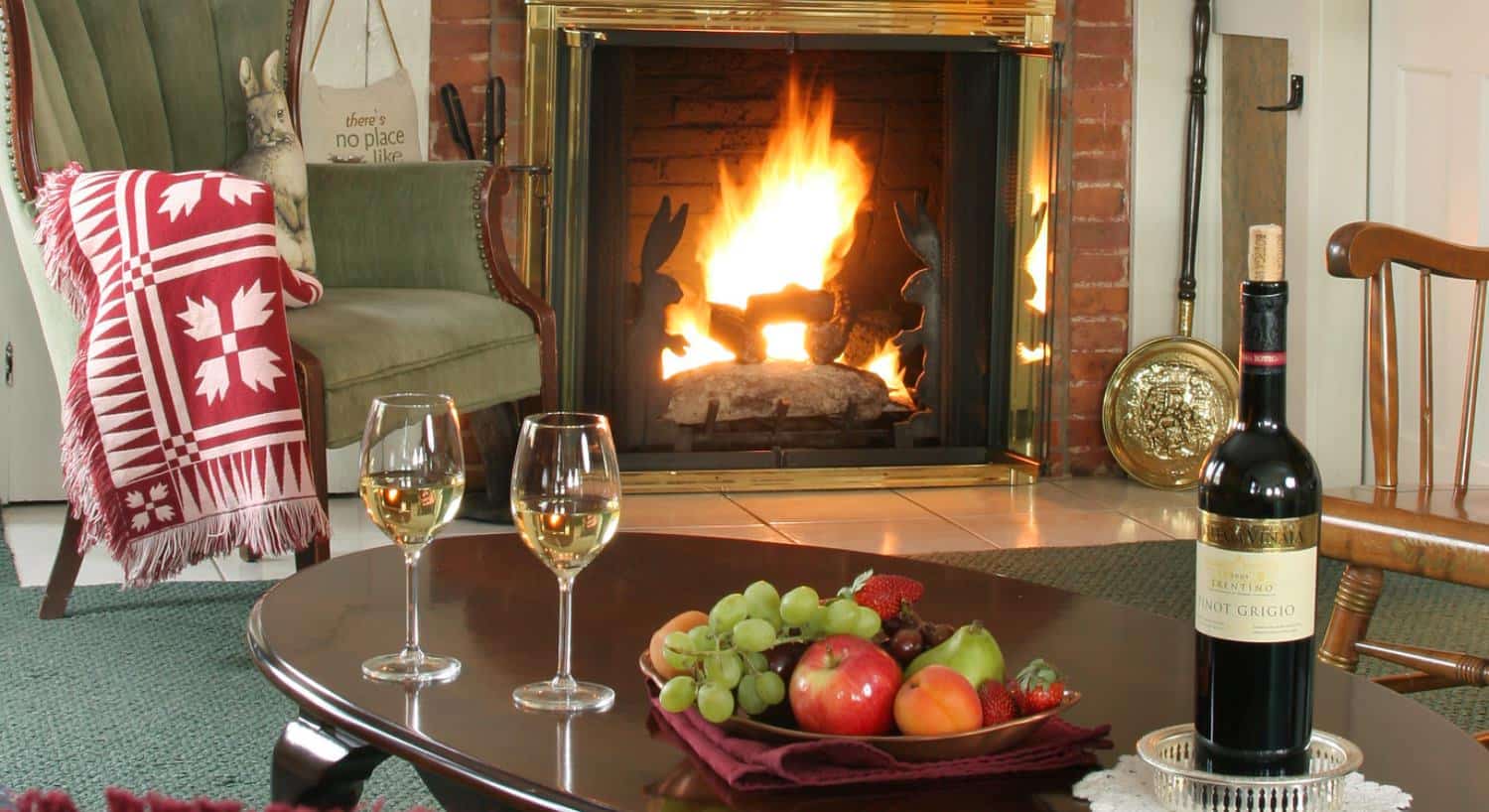 Close up view of white wine in glasses and bowl of fruit on dark wooden coffee table near an antique wooden armchair and fireplace