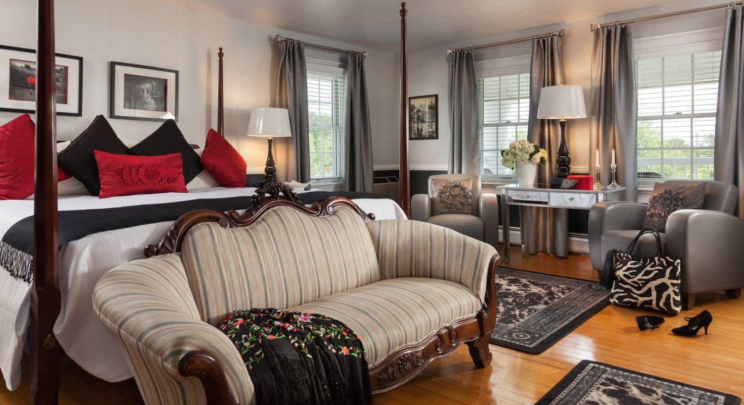 Large bedroom with hardwood flooring, dark hardwood four-poster bed, white bedding, black and red pillows, antique wooden couch, and sitting area with gray leather chairs