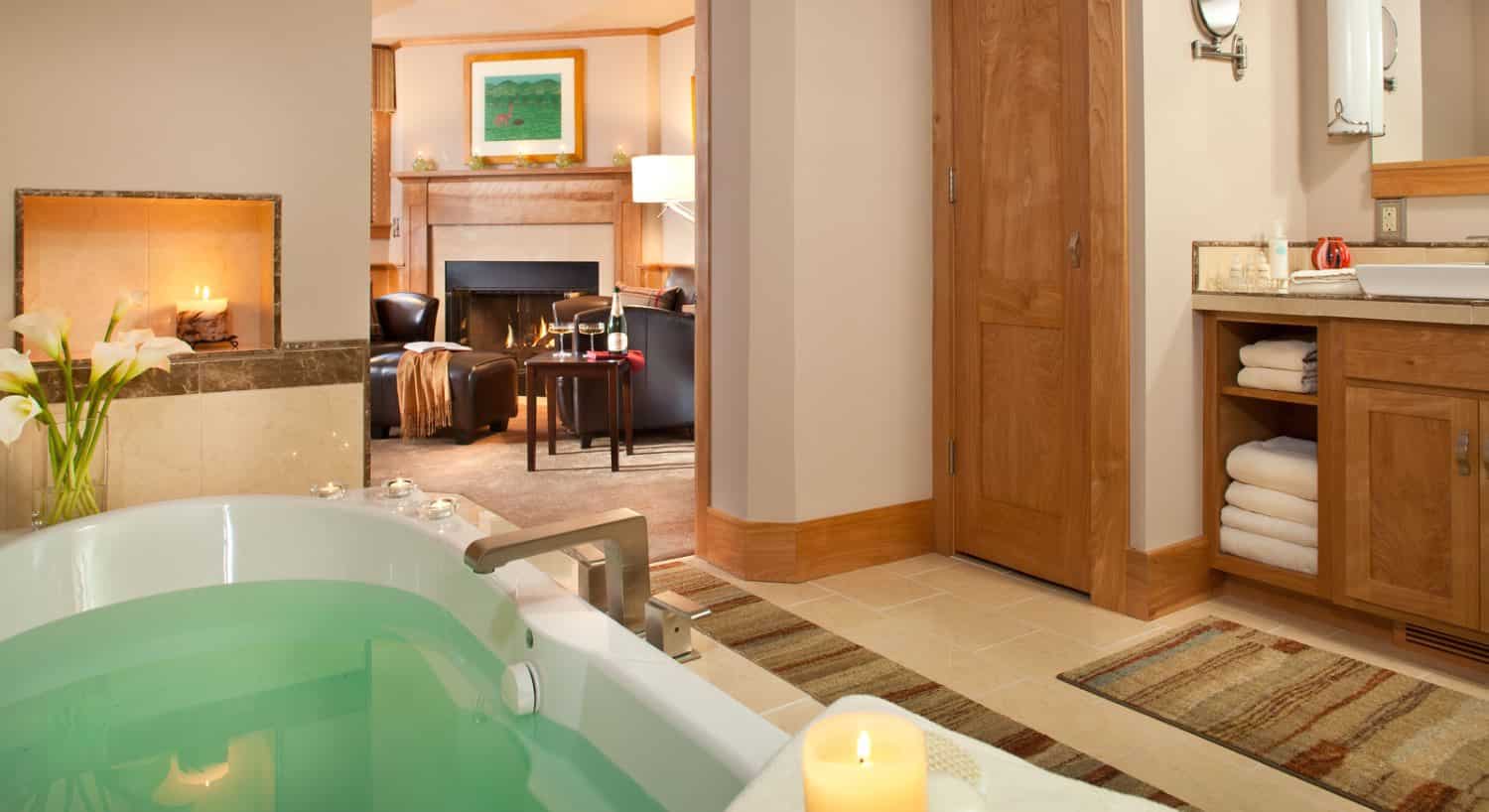 Large whirlpool tub filled with water surrounded by candles and flowers with view into bedroom with sitting area and fireplace