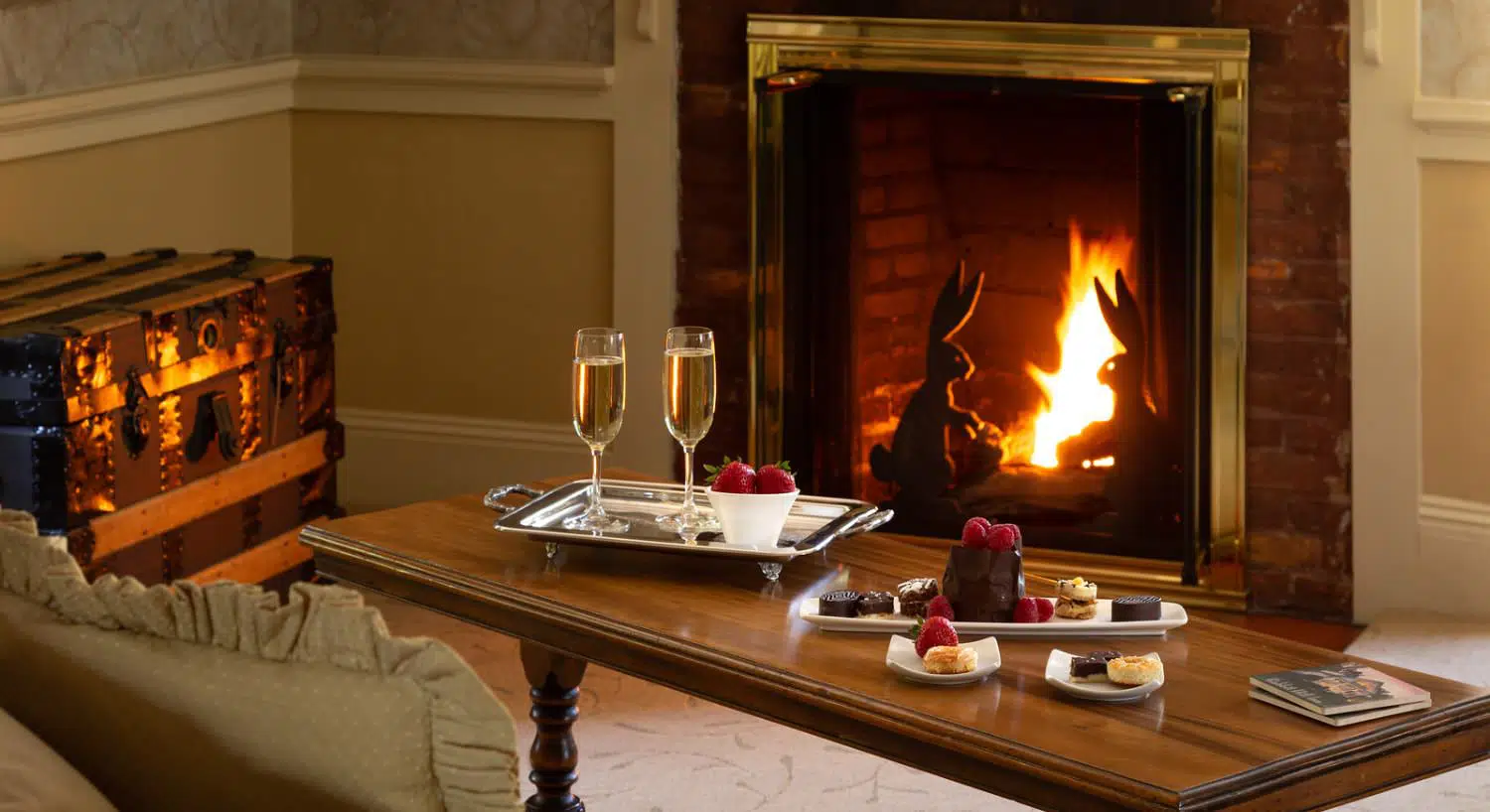 Close up view of sitting area with dark wooden coffee table, silver tray with champagne glasses and bowl of strawberries, white tray with desserts, and a fireplace