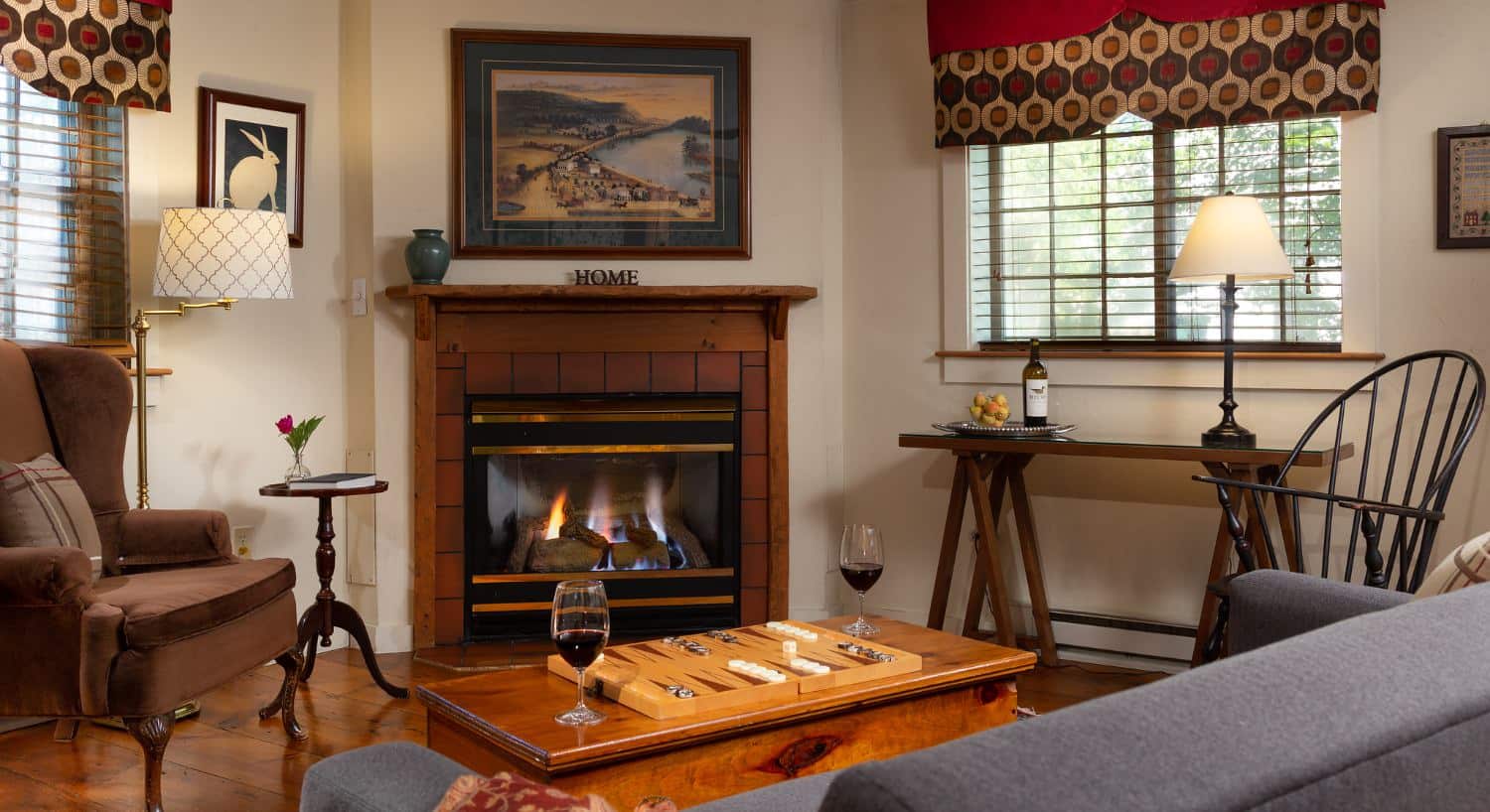 Bedroom's sitting area with hardwood floors, cream walls, gray upholstered couch, brown upholstered arm chair, wooden trunk as a coffee table, and fireplace