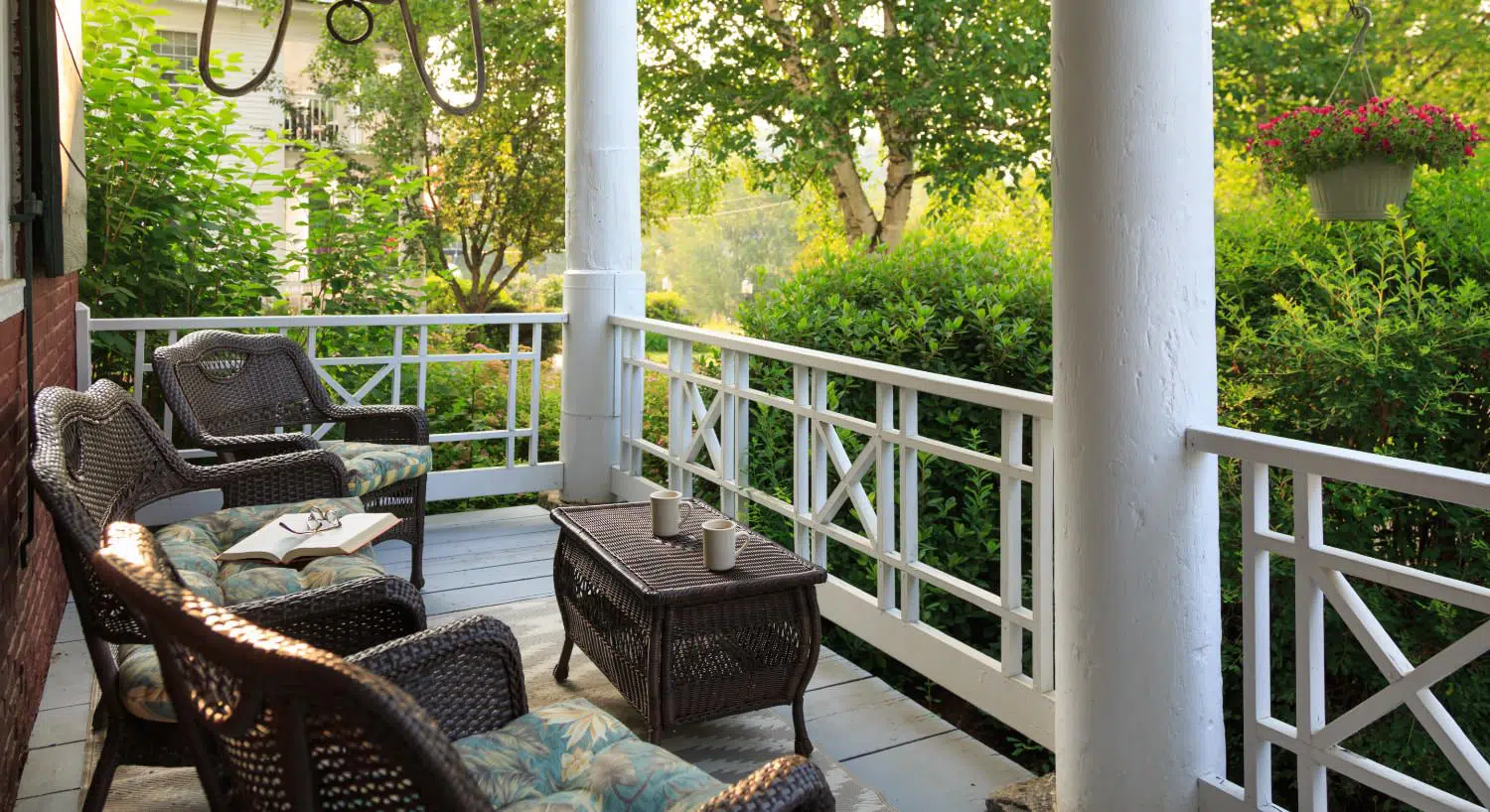 Front porch with dark brown wicker patio furniture, floral cushions, and view of green bushes and trees