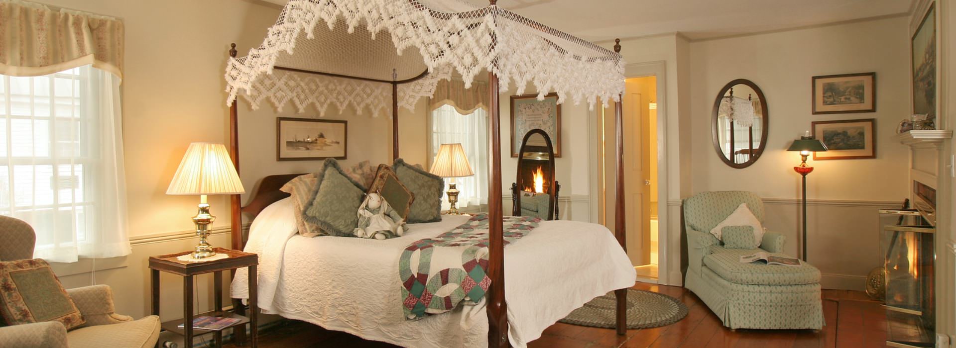 Large bedroom suite with hardwood floors, light-colored walls, dark wooden four-poster canopy bed with white bedding, sitting area, and fireplace