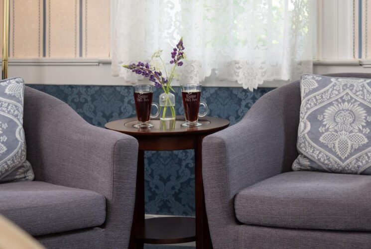 Sitting area with gray upholstered arm chairs, dark wooden table, two glasses filled with a dark drink, and sprigs of lavendar in glass vase