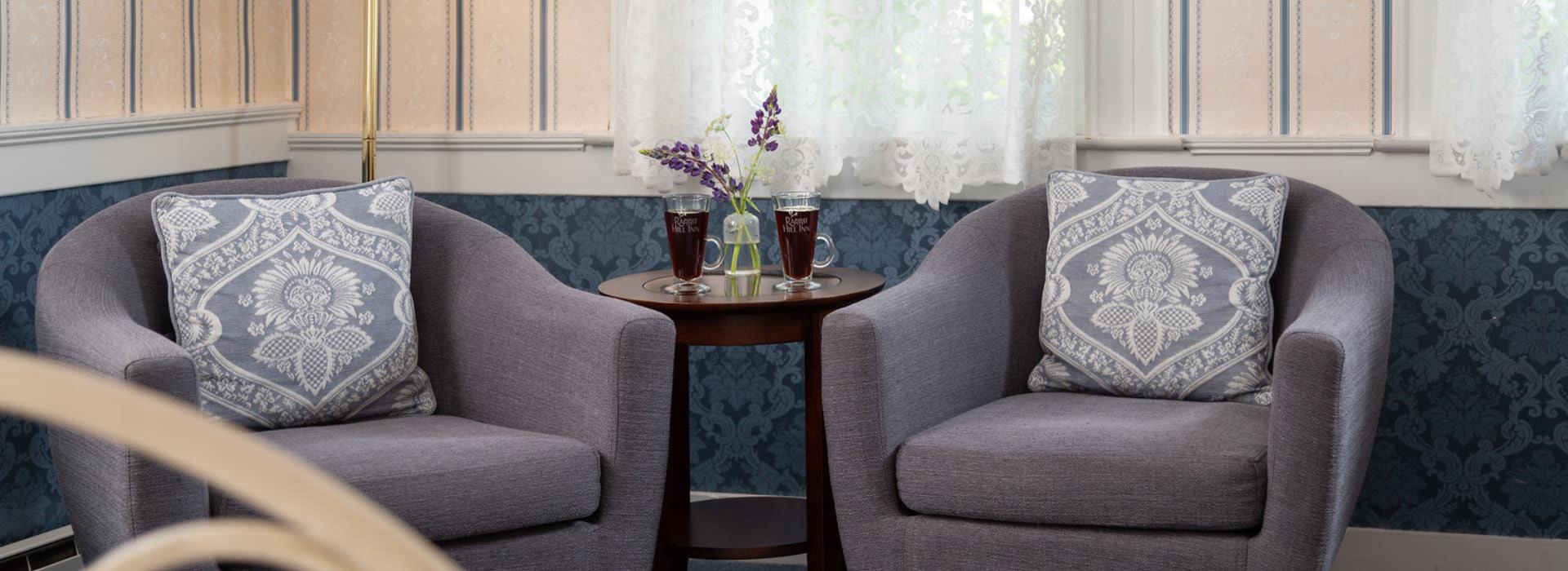 Sitting area with gray upholstered arm chairs, dark wooden table, two glasses filled with a dark drink, and sprigs of lavendar in glass vase