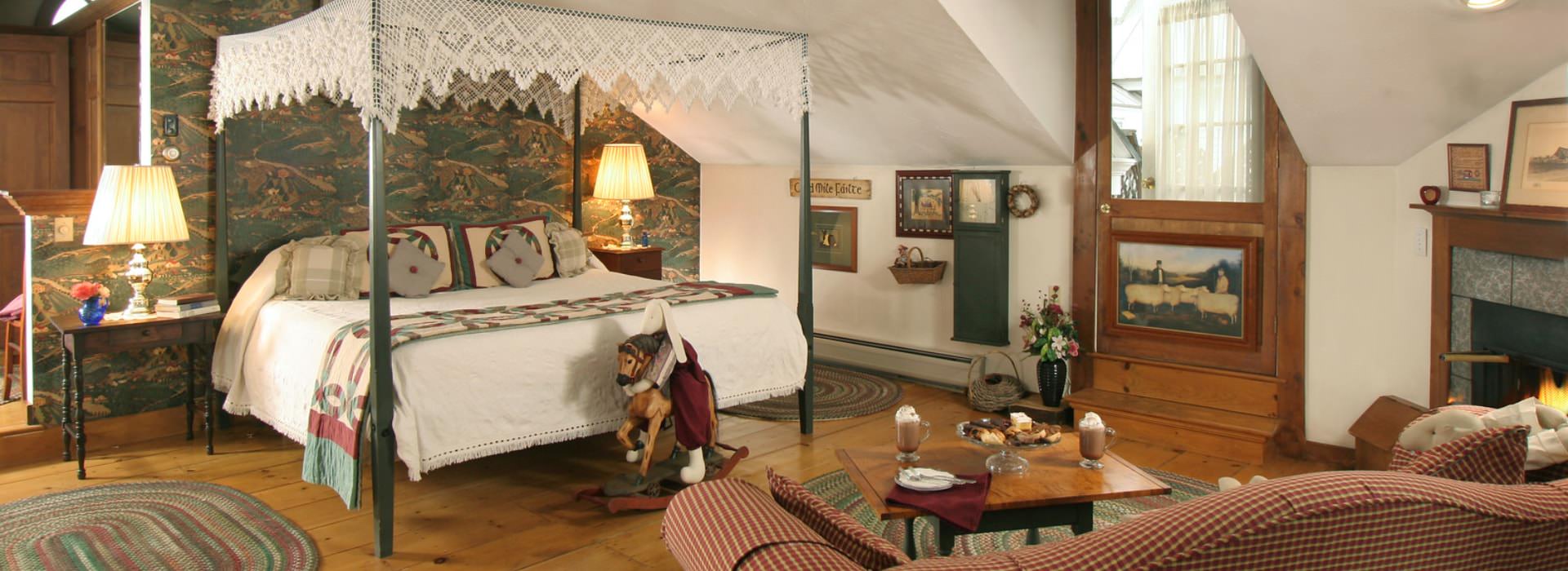 Large bedroom suite with hardwood floors, cream walls, custom wallpaper, dark green wooden four-poster bed, white bedding, sitting area, and fireplace