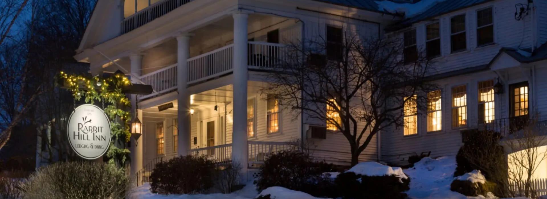 Exterior view of the property painted white with front porch and second level porch all surrounded by snow and winter-looking trees and shrubs at night