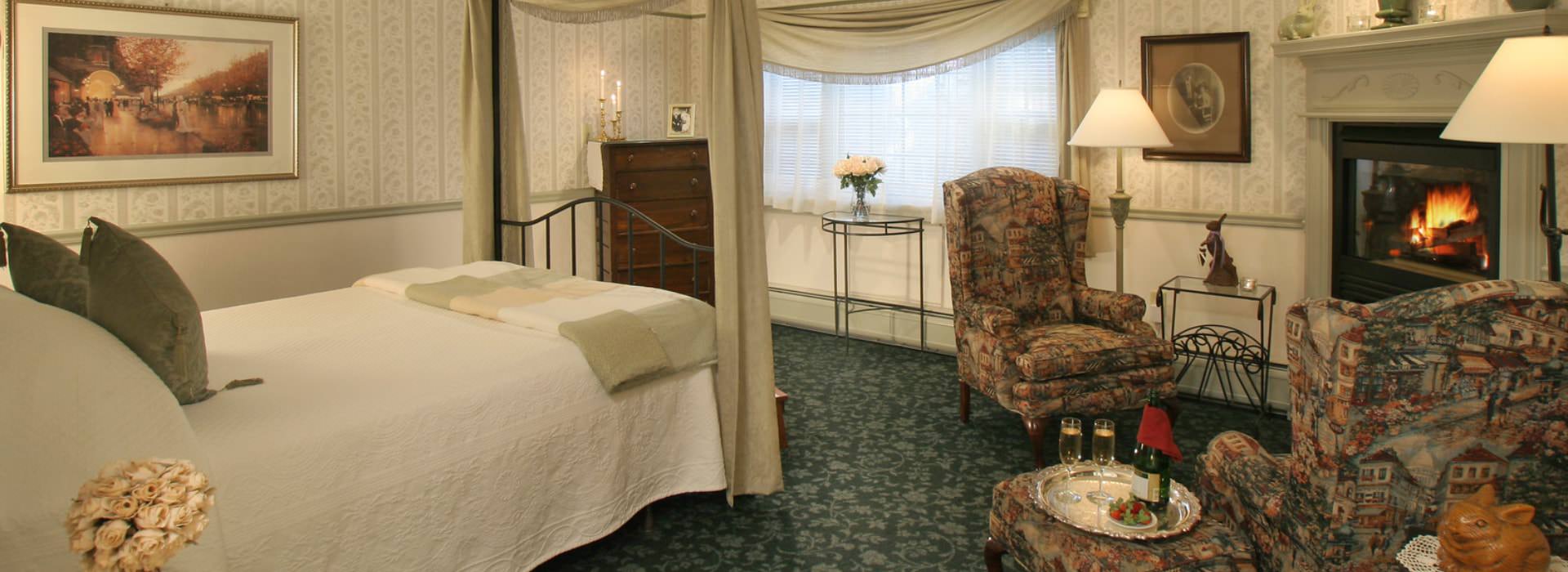 Large bedroom suite with wrought iron canopy bed, white bedding, dark green floral carpeting, sitting area, and fireplace