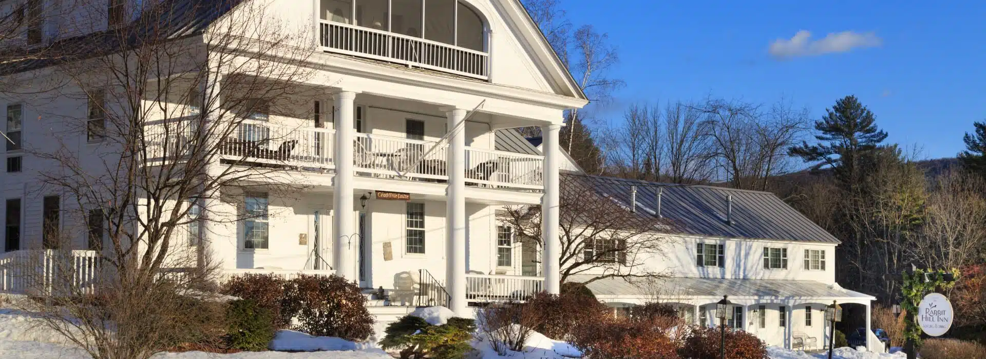 Exterior view of the property painted white with front porch and second level porch all surrounded by snow and winter-looking trees and shrubs with blue clouds in the sky