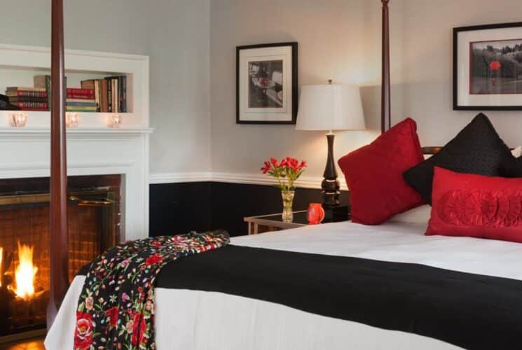 Bedroom with light-gray walls on top and black on bottom of chair rail, dark wooden four-poster bed, white bedding, black and red pillows, and a fireplace