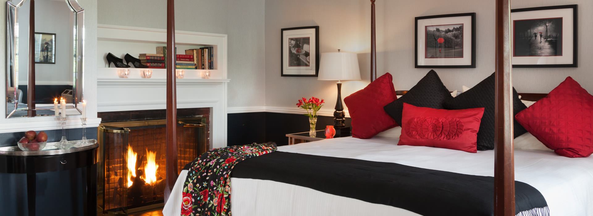 Bedroom with light-gray walls on top and black on bottom of chair rail, dark wooden four-poster bed, white bedding, black and red pillows, and a fireplace