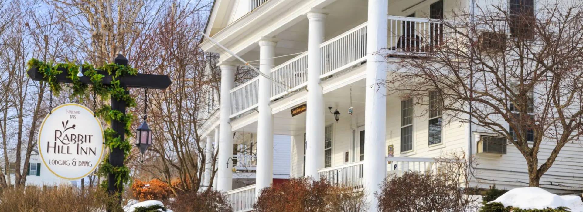 Exterior view of the property painted white with front porch and second level porch all surrounded by snow and winter-looking trees and shrubs