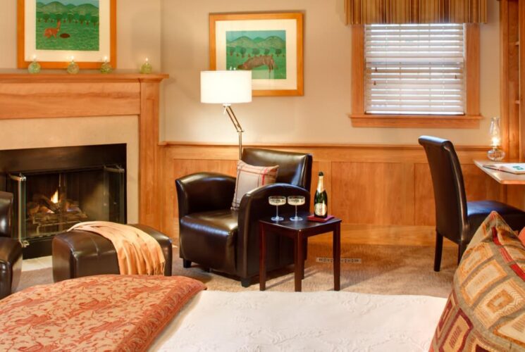 Large bedroom with cream colored walls, wood paneled chair railing, fireplace, and sitting area with dark brown leather chairs