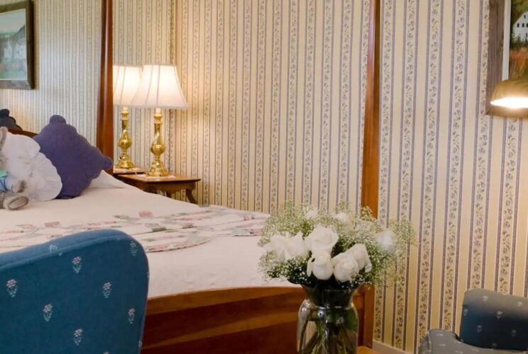 Bedroom with cream and floral striped wallpaper, wooden four-poster bed, white bedding, purple pillows, and sitting area