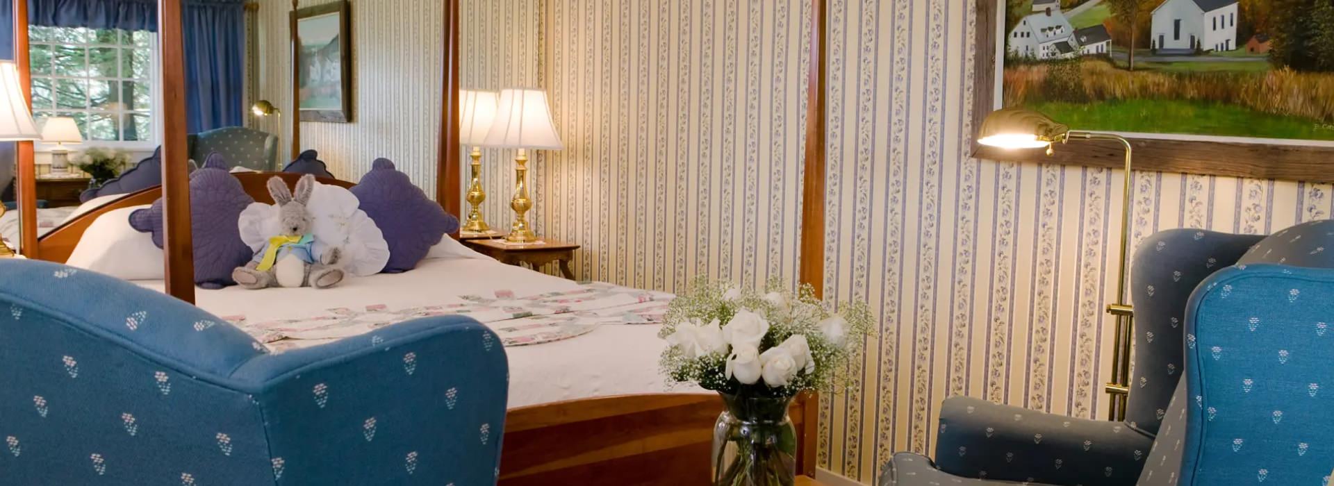 Bedroom with cream and floral striped wallpaper, wooden four-poster bed, white bedding, purple pillows, and sitting area