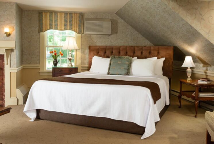 Large bedroom suite with light-colored carpet, fillagree wallpaper, light-tan wall under chair rail, brown upholstered headboard, white bedding, sitting area, and fireplace