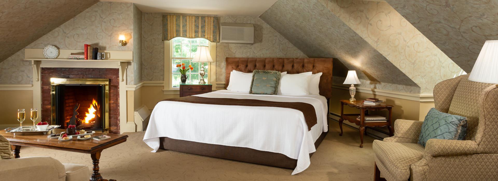 Large bedroom suite with light-colored carpet, fillagree wallpaper, light-tan wall under chair rail, brown upholstered headboard, white bedding, sitting area, and fireplace