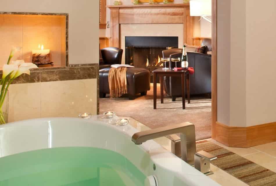 Large whirlpool tub filled with water surrounded by candles and flowers with view into bedroom with sitting area and fireplace