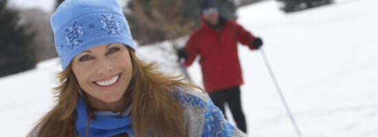 Woman smiling as she enjoys cross country skiing and a man standing in the background|a couple snowshoeing in the snowy woods|