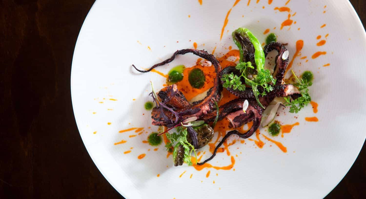 Close up view of cooked octopus decorated with roasted peppers and cilantro on a white porcelain plate